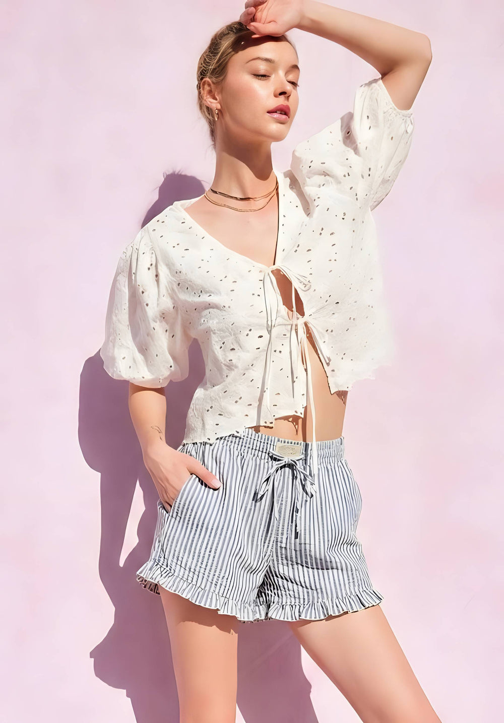 A woman modeling a white eyelet short-sleeve crop top with a front tie closure and striped blue-and-white ruffled shorts, posed against a light pink wall with strong sunlight and shadows.