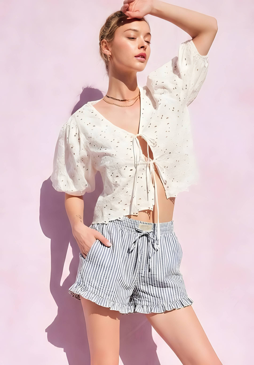 A woman modeling a white eyelet short-sleeve crop top with a front tie closure and striped blue-and-white ruffled shorts, posed against a light pink wall with strong sunlight and shadows.