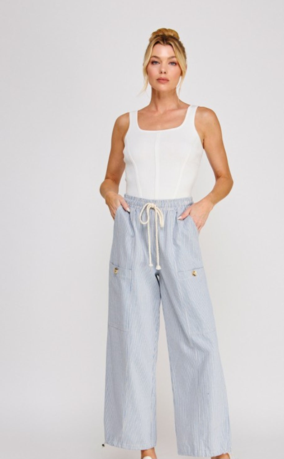 Woman wearing a white sleeveless top and light blue pants with drawstring on a plain background