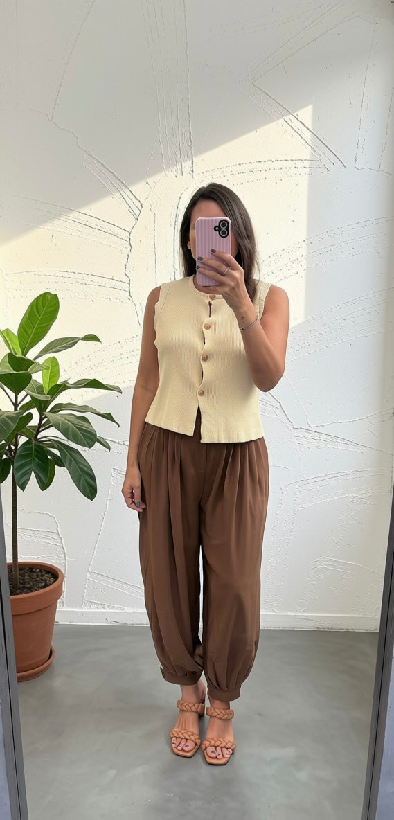 A woman takes a mirror selfie showing a two-piece outfit consisting of a sleeveless cream button-front vest and brown harem pants, standing in a room with a white wall and a potted plant.