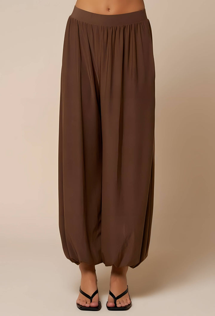 A person wearing loose, draped harem-style pants in a medium brown color, paired with black thong sandals, shown against a plain beige studio background.