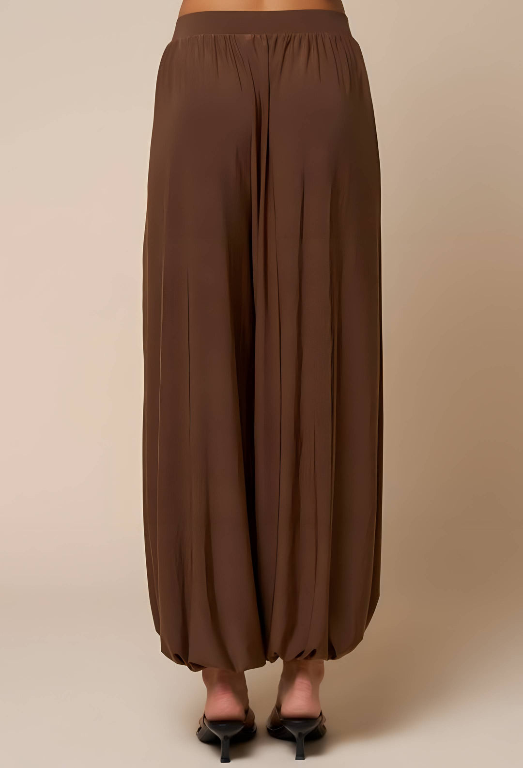 A person shown from the back wearing loose, wide-leg brown pants with a gathered elastic waistband and black pointed heels, against a plain beige studio background.