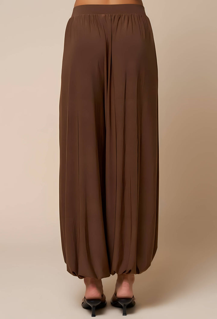 A person shown from the back wearing loose, wide-leg brown pants with a gathered elastic waistband and black pointed heels, against a plain beige studio background.
