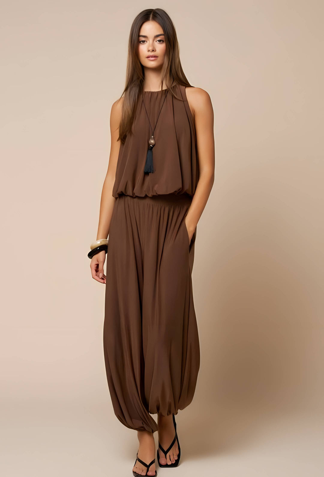 A woman modeling a sleeveless brown two-piece outfit consisting of a draped halter-style top and matching loose harem-style pants, photographed against a plain beige studio background.