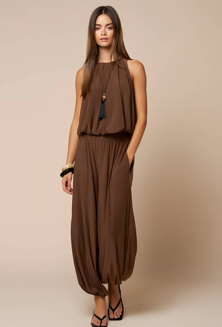 A woman modeling a sleeveless brown two-piece outfit consisting of a draped halter-style top and matching loose harem-style pants, photographed against a plain beige studio background.