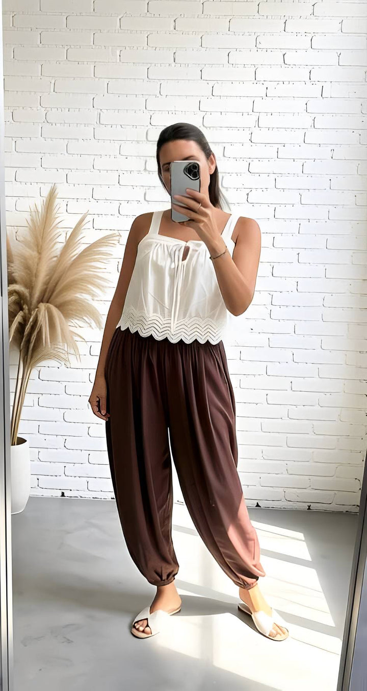 A woman takes a mirror selfie wearing a white sleeveless top with a scalloped hem and loose brown harem-style pants, standing in front of a white brick wall.