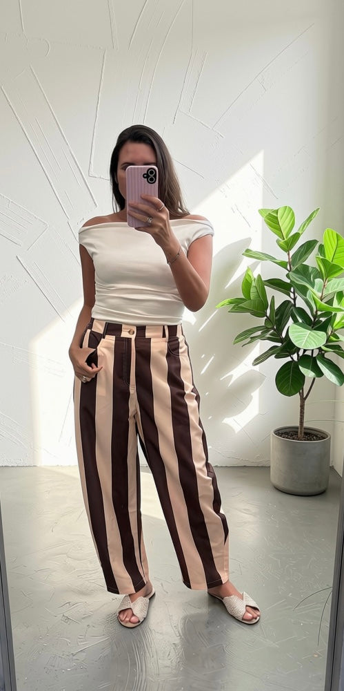 A woman takes a mirror selfie wearing a white off-the-shoulder top and high-waisted, wide-leg trousers with vertical dark brown and beige stripes. She is standing in a bright room with a potted plant on the right.