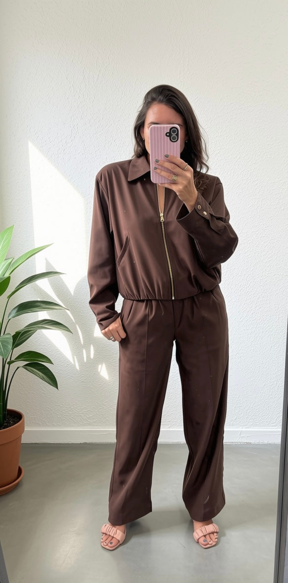 A woman takes a mirror selfie wearing a matching brown long-sleeve collared zip-up jacket and wide-leg pants set, standing in a room with a potted plant on the left.