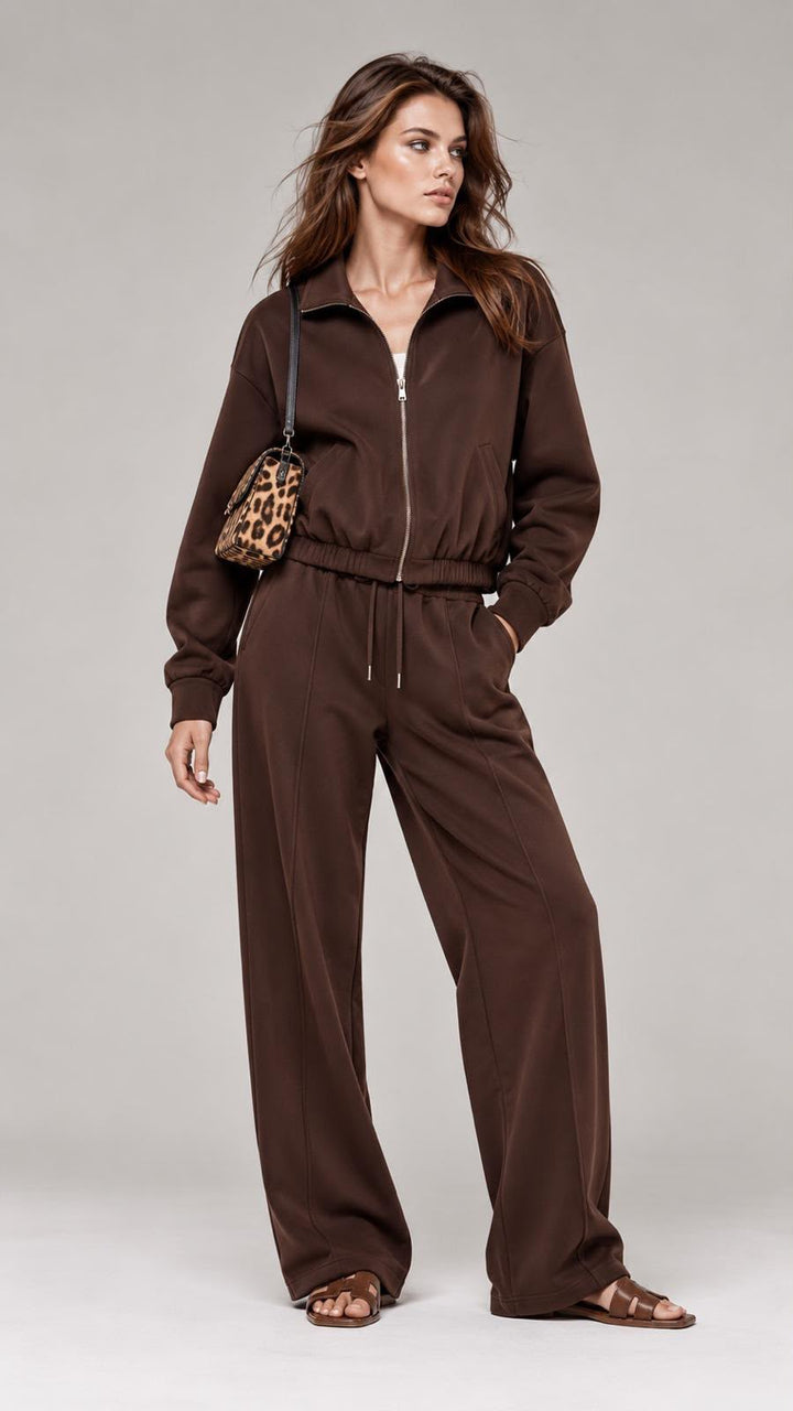 A woman modeling a dark brown zip-up sweatshirt and matching wide-leg sweatpants, styled with a leopard-print crossbody bag and brown flat sandals against a light gray studio background.