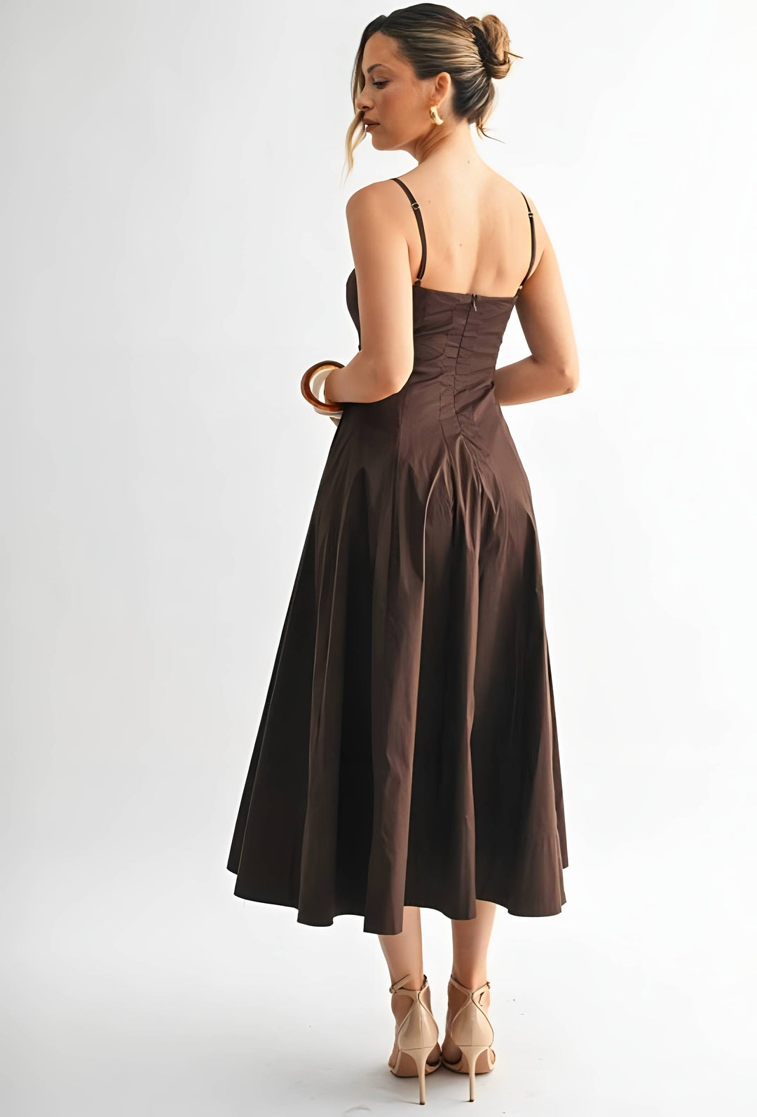 A woman modeling a dark brown midi dress from the back, featuring thin spaghetti straps, a structured corset-style back, and a flared skirt, set against a plain white studio background.
