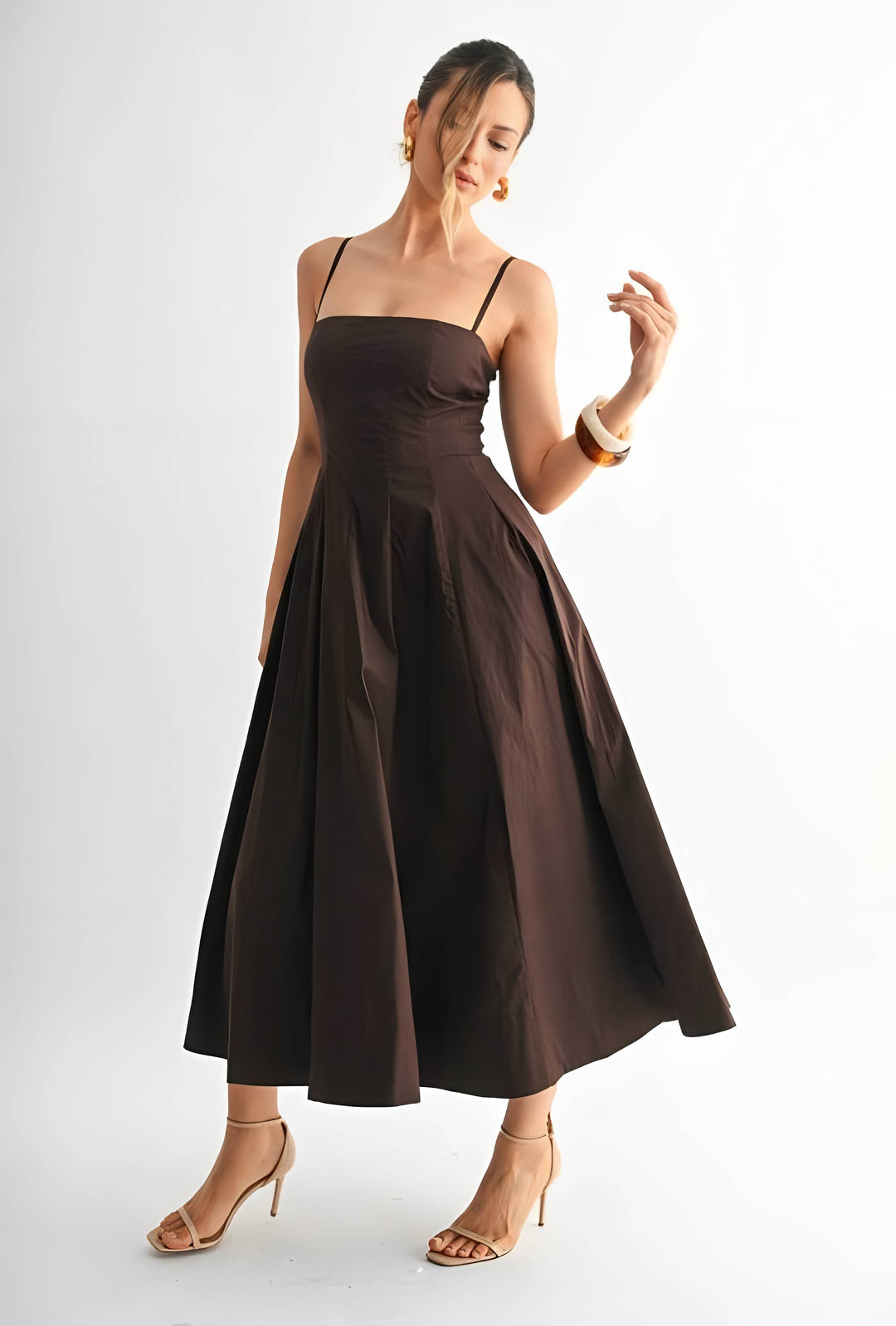A woman modeling a dark brown midi dress with thin spaghetti straps, a structured square neckline, a fitted waist, and a full flared skirt, paired with nude high-heeled sandals against a plain white studio background.