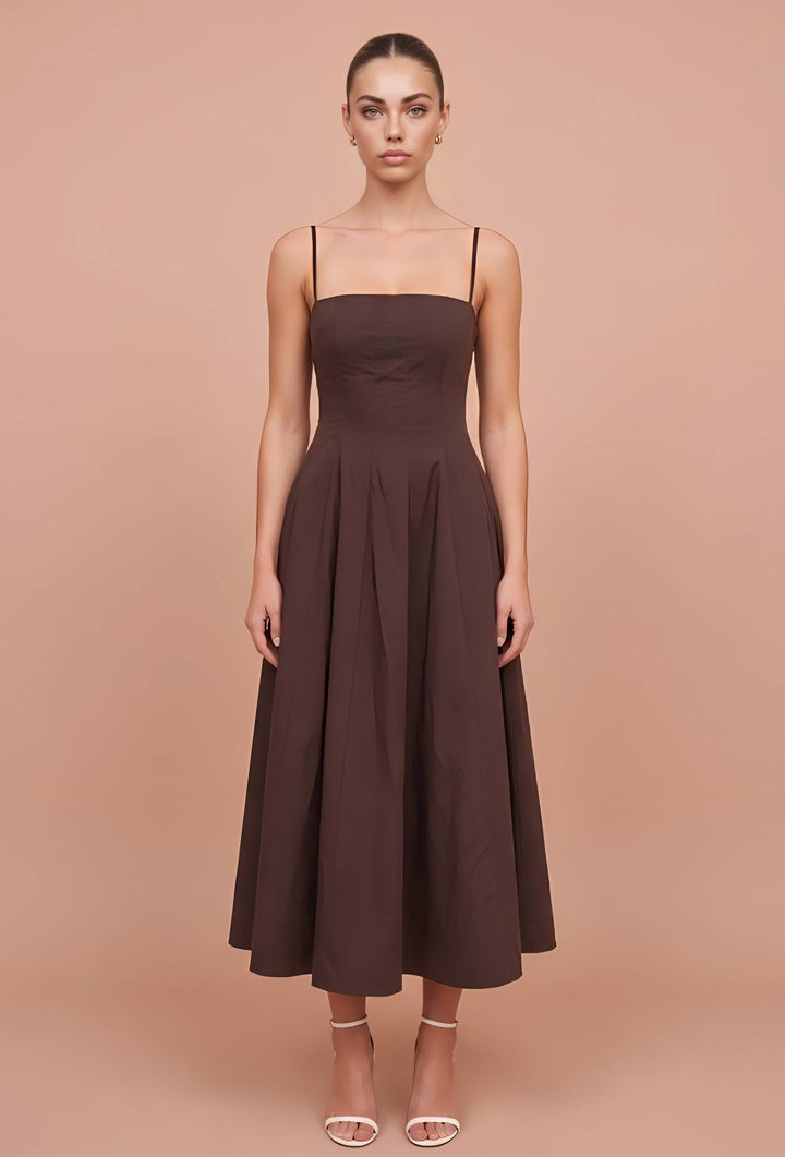 A woman modeling a dark brown midi dress with thin spaghetti straps, a square neckline, a structured bodice, and a flared skirt, set against a solid peach studio background.
