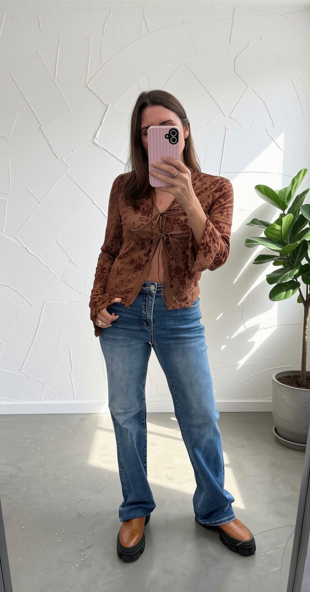 A woman takes a mirror selfie wearing a brown patterned long-sleeve cropped top, blue flare jeans, and brown shoes in a bright room with a potted plant.
