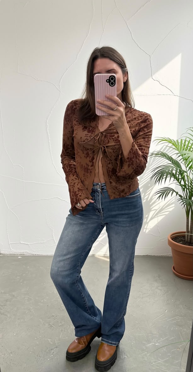 A woman takes a mirror selfie wearing a brown patterned long-sleeve tie-front top, blue jeans, and brown leather boots, standing in a room with a white wall and a potted plant.
