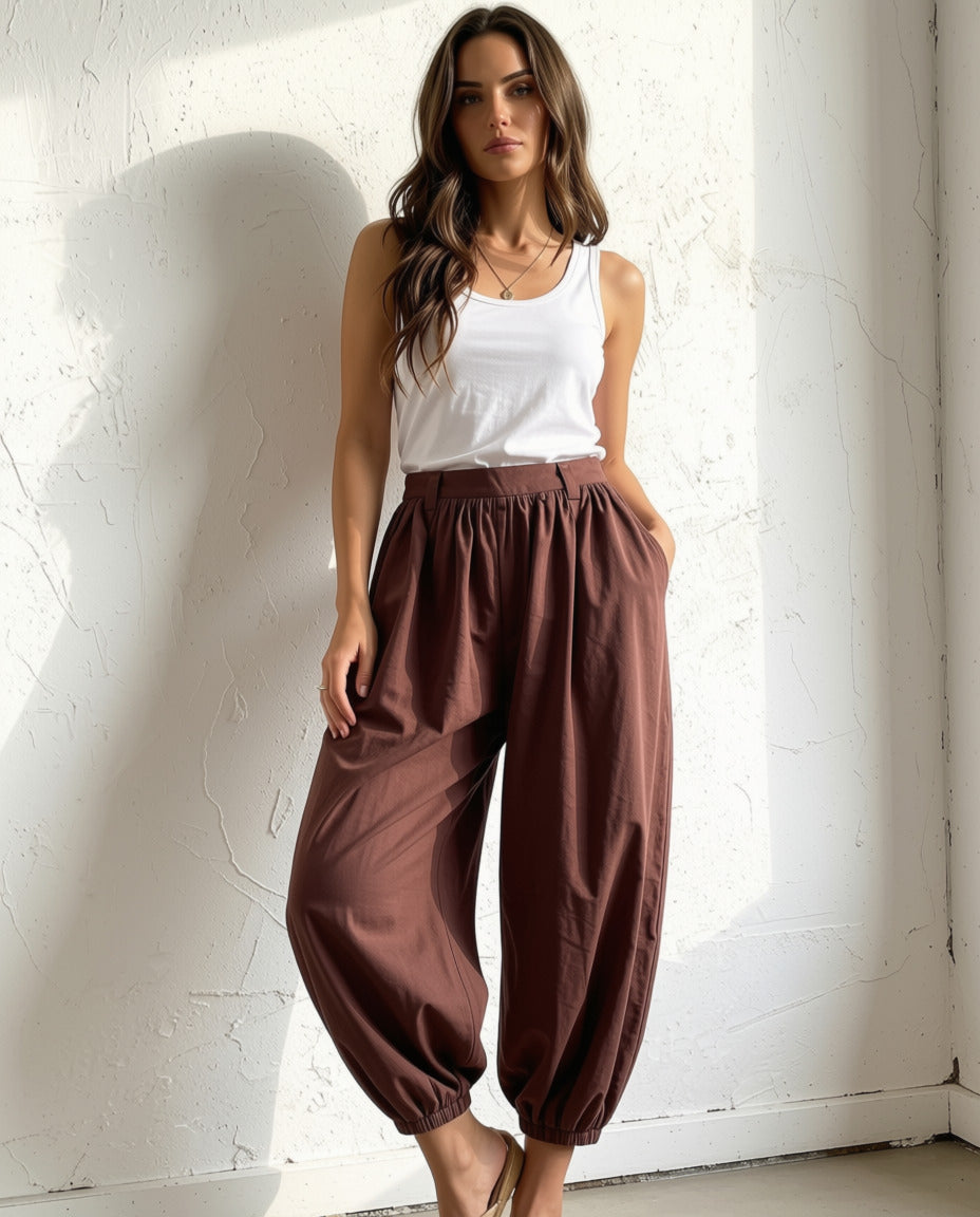 A woman modeling loose brown harem-style pants with an elastic waist and gathered cuffs, paired with a white tank top, standing against a textured white wall.