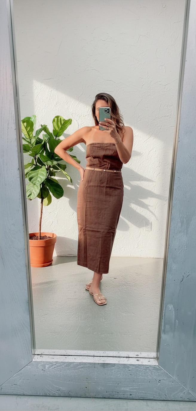 Woman taking a mirror selfie wearing a brown strapless top and skirt with a plant in the background.