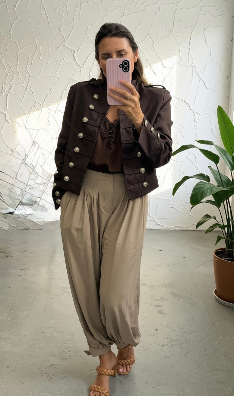 A woman takes a mirror selfie showing an outfit consisting of a dark brown double-breasted jacket with silver buttons, a brown lace-up top, beige wide-leg pants, and tan sandals.