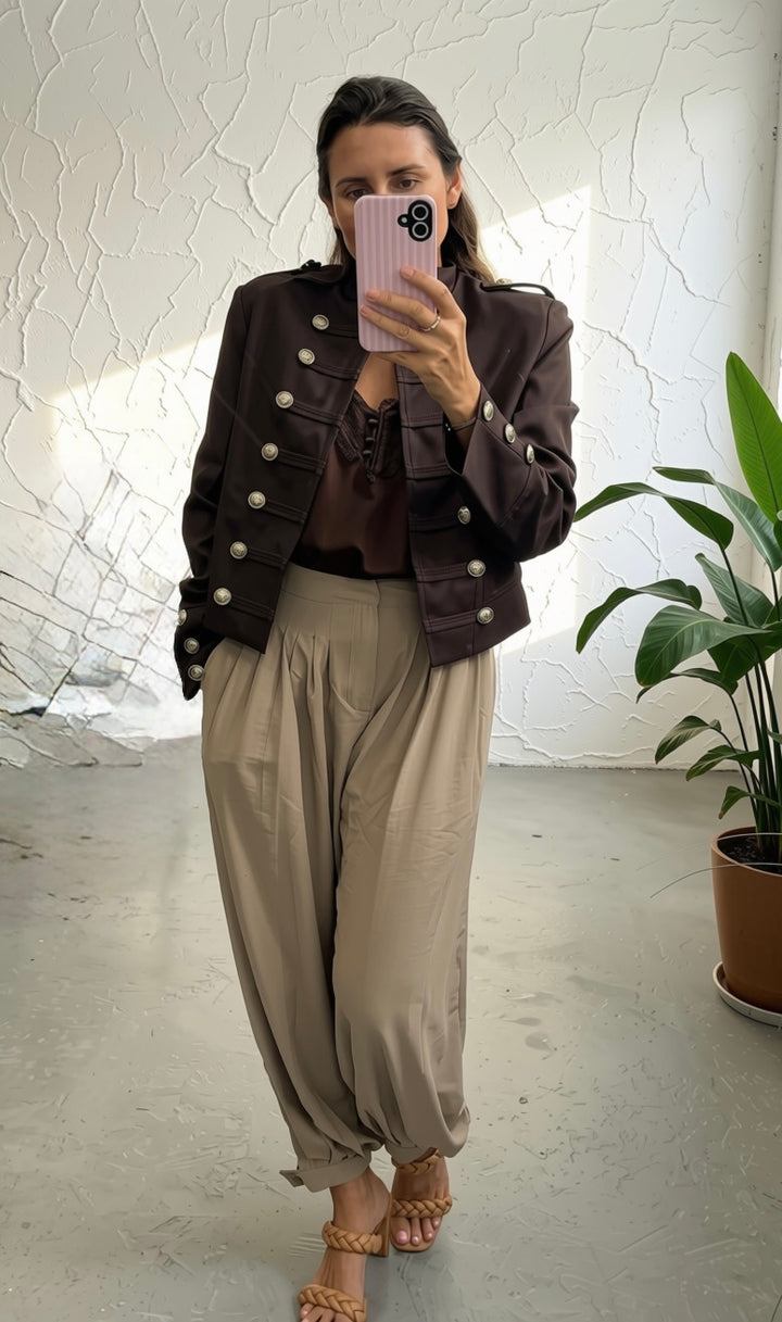 A woman takes a mirror selfie showing an outfit consisting of a dark brown double-breasted jacket with silver buttons, a brown lace-up top, beige wide-leg pants, and tan sandals.