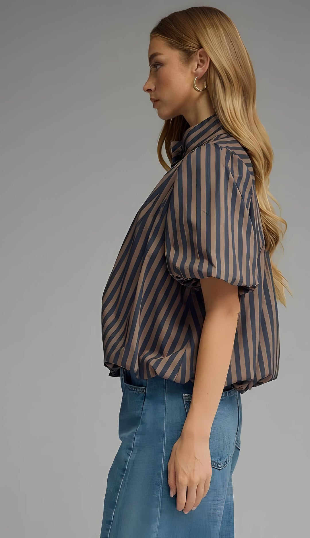A woman shown in side profile wearing a short-sleeve button-up shirt with vertical brown and navy stripes, paired with blue jeans, against a solid gray studio background.
