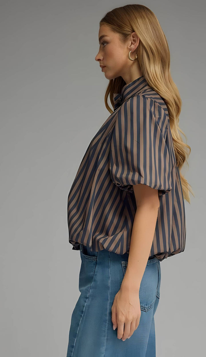 A woman shown in side profile wearing a short-sleeve button-up shirt with vertical brown and navy stripes, paired with blue jeans, against a solid gray studio background.