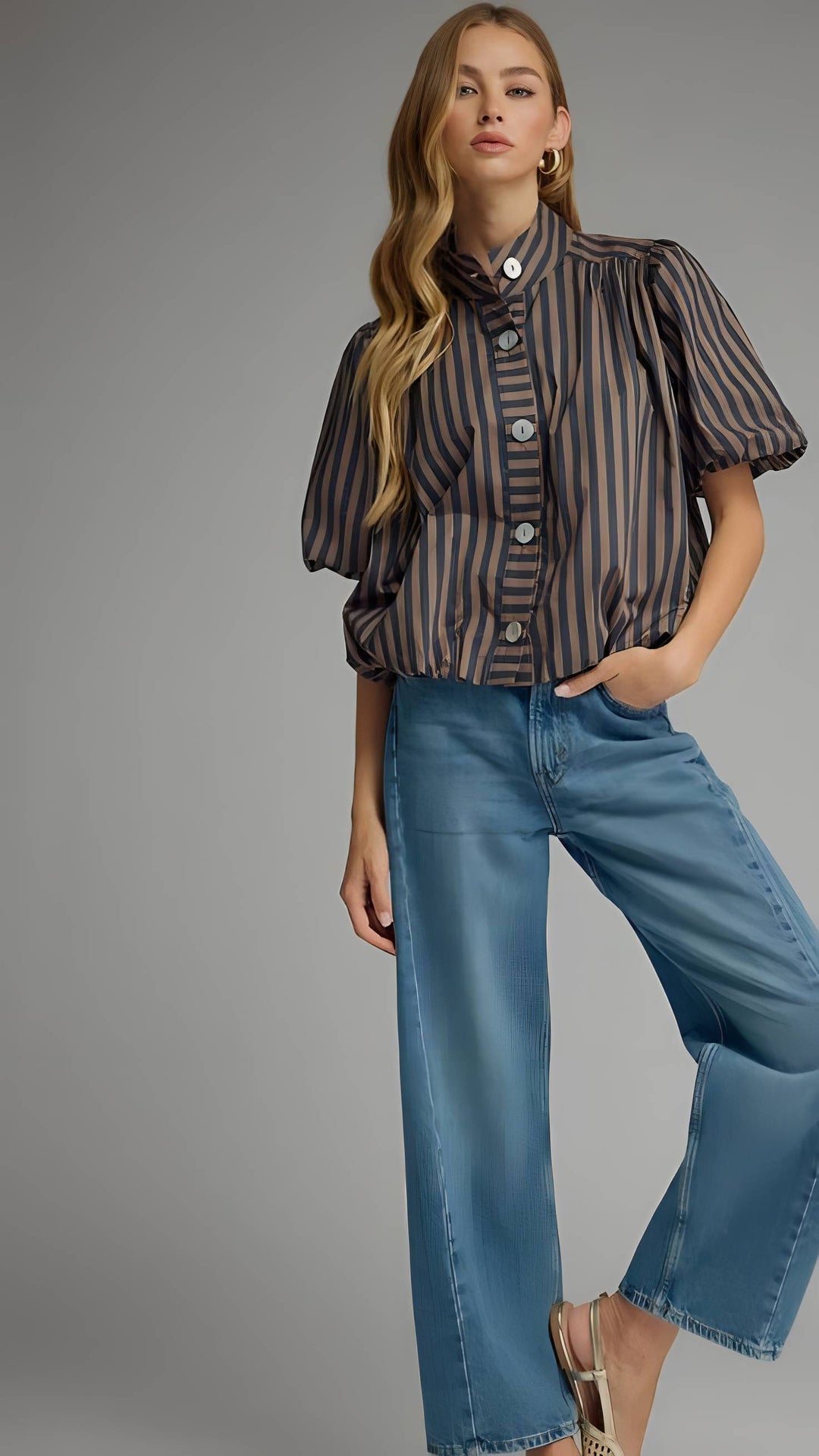 A woman modeling a short-sleeve button-front blouse with vertical dark brown and beige stripes, paired with wide-leg blue jeans, against a plain gray studio background.