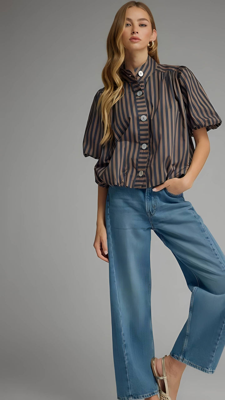 A woman modeling a short-sleeve button-front blouse with vertical dark brown and beige stripes, paired with wide-leg blue jeans, against a plain gray studio background.