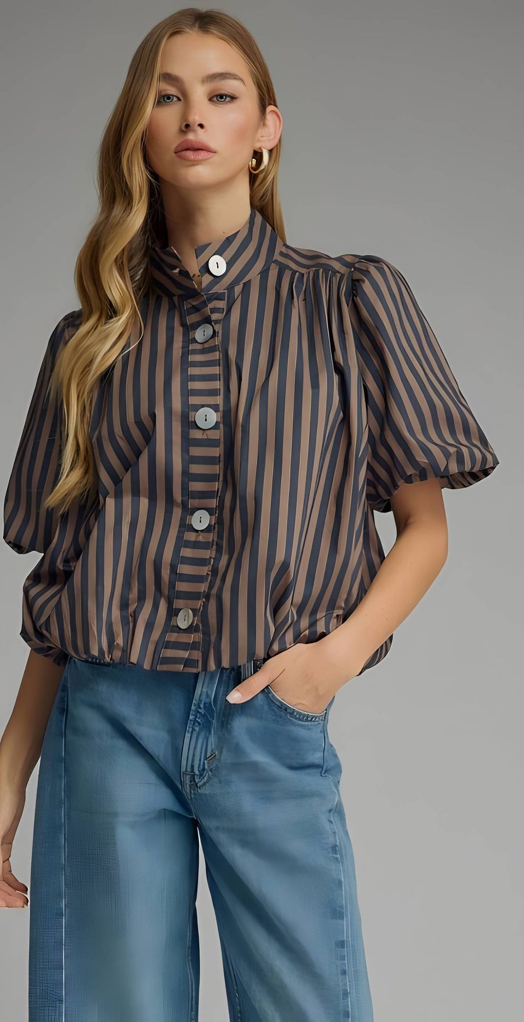 A woman modeling a short-sleeve button-front blouse with vertical dark brown and beige stripes, paired with wide-leg blue jeans, against a plain gray studio background.