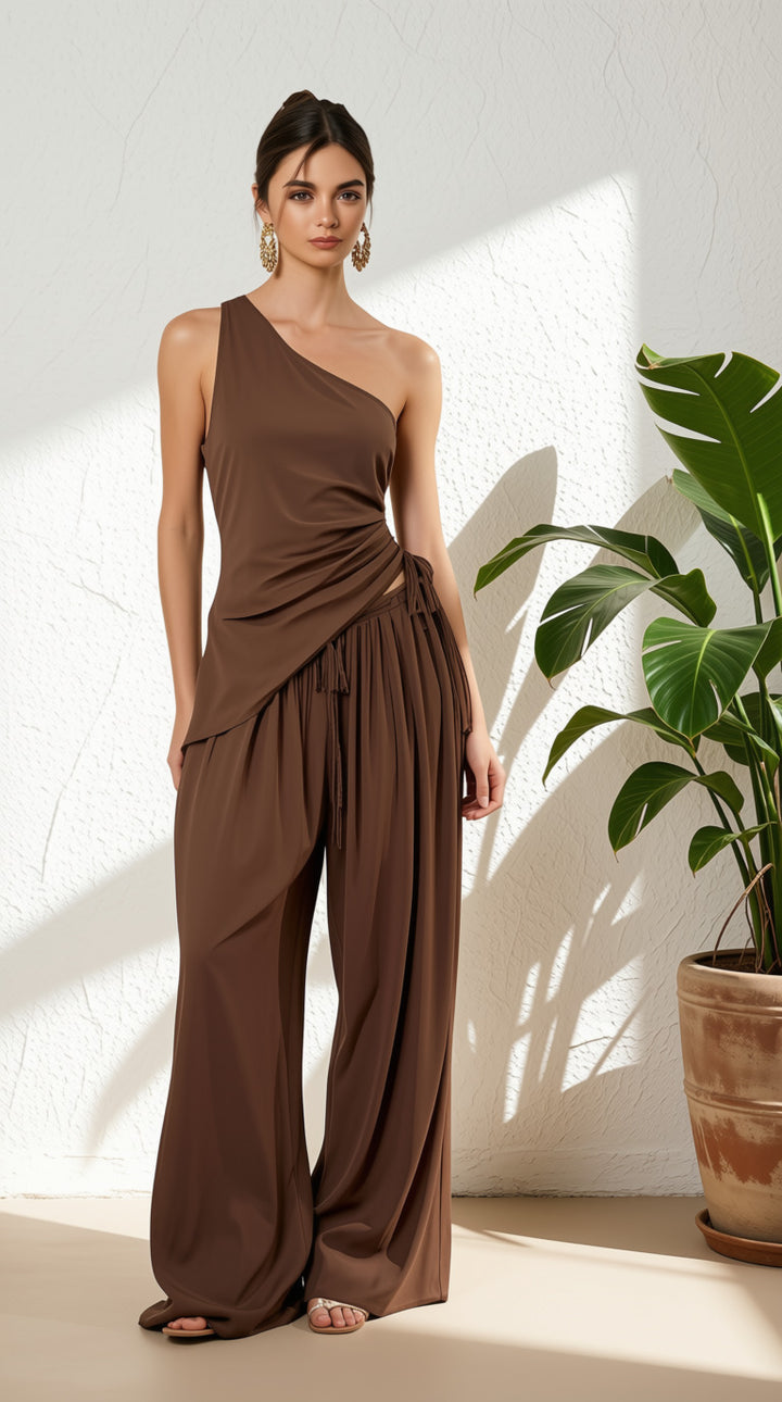 Woman wearing a 2 piece pant set with a one shoulder top and baggy pants in front of a white wall.