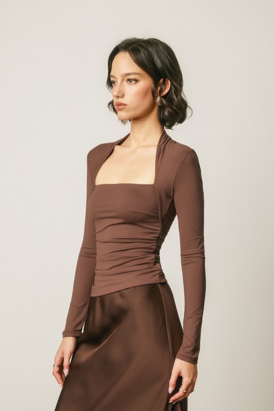 A woman modeling a brown long-sleeve top with a square neckline and gathered waist, paired with a matching brown satin skirt, against a light gray studio background.