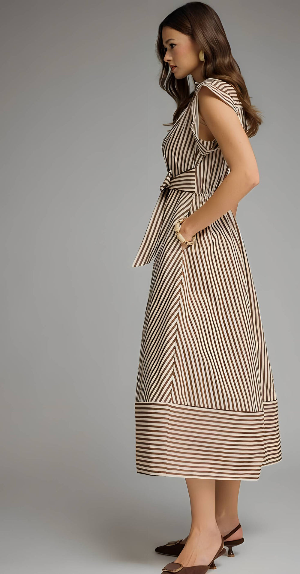 A woman modeling a sleeveless midi dress with vertical brown and beige stripes, featuring a tie waist and side pockets, photographed against a neutral gray studio background.
