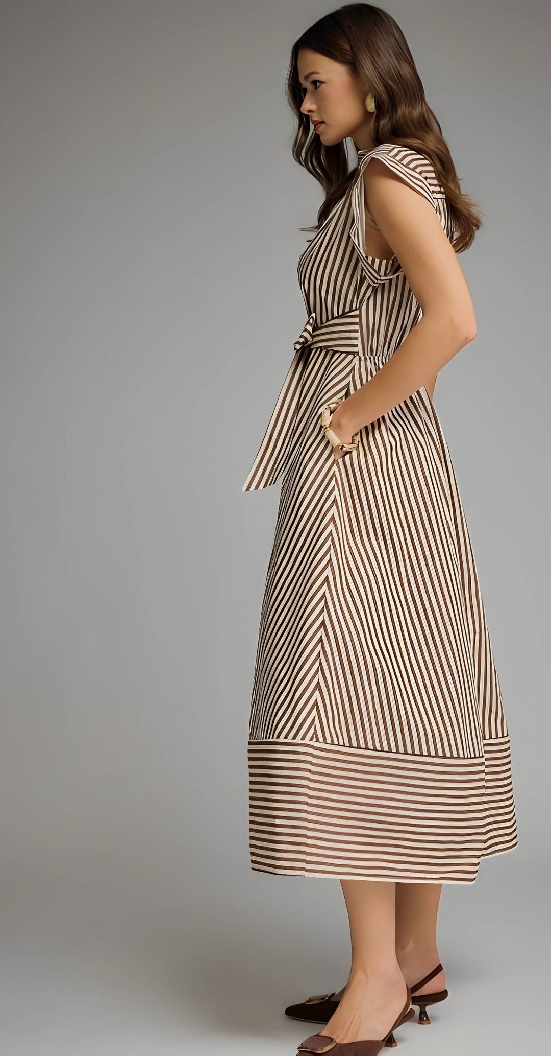 A woman modeling a sleeveless midi dress with vertical brown and beige stripes, featuring a tie waist and side pockets, photographed against a neutral gray studio background.