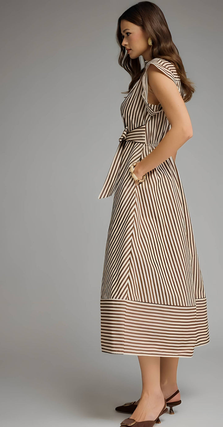 A woman modeling a sleeveless midi dress with vertical brown and beige stripes, featuring a tie waist and side pockets, photographed against a neutral gray studio background.