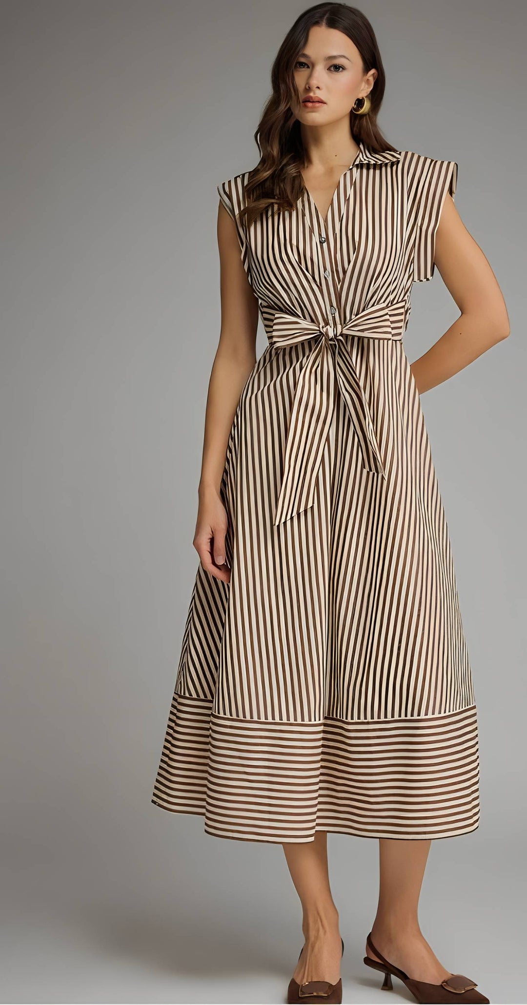 A woman models a sleeveless midi dress with vertical brown and beige stripes, featuring a tie-front waist and a tiered skirt, set against a solid gray studio background.