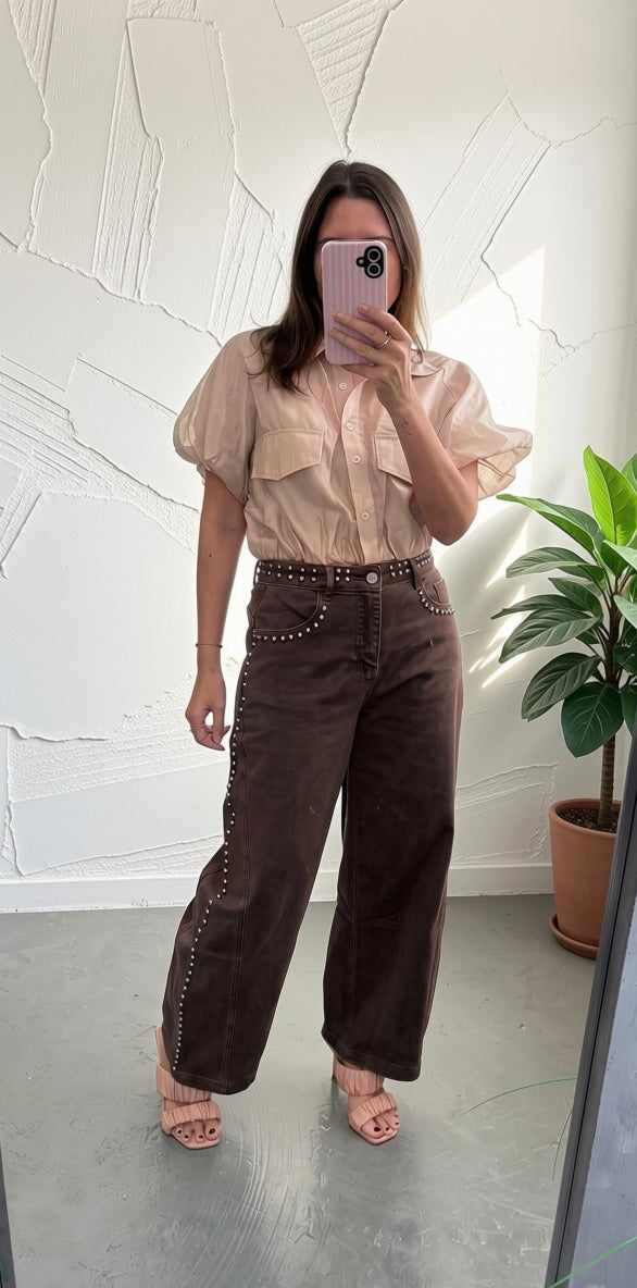 A woman takes a mirror selfie wearing a beige short-sleeve button-up shirt with puff sleeves and dark brown wide-leg corduroy pants featuring silver stud detailing along the waistband and leg hems.