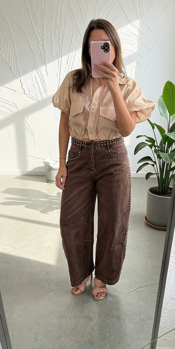 A woman takes a mirror selfie showing a two-piece outfit consisting of a beige puff-sleeve button-up shirt and brown wide-leg pants with silver studs on the waistband and sides. She is standing in a bright room with a potted plant in the background.