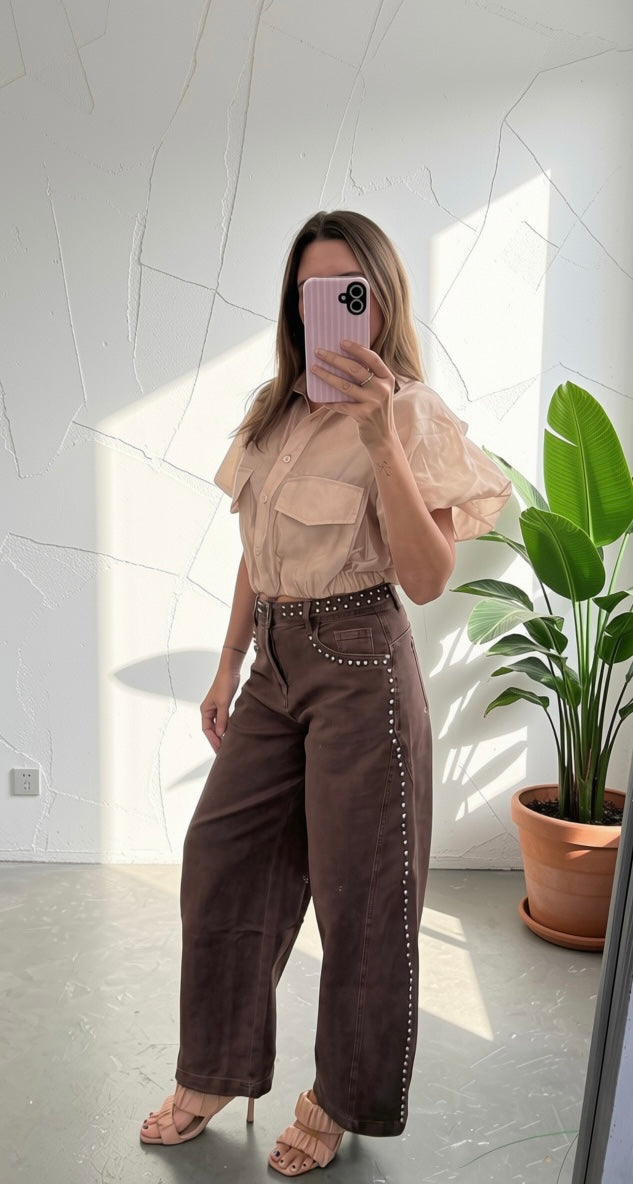 A woman takes a mirror selfie in a room with bright sunlight and shadows, wearing a light beige ruffled short-sleeve blouse and dark brown wide-leg trousers with silver studs.
