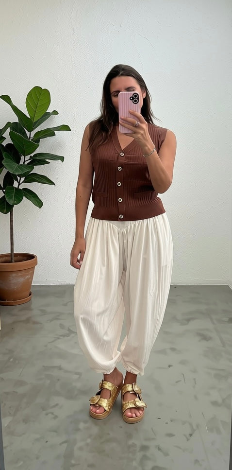 A woman takes a mirror selfie wearing a brown ribbed sleeveless button-front top and cream harem-style pants, paired with gold sandals, in a minimalist room with a potted plant.