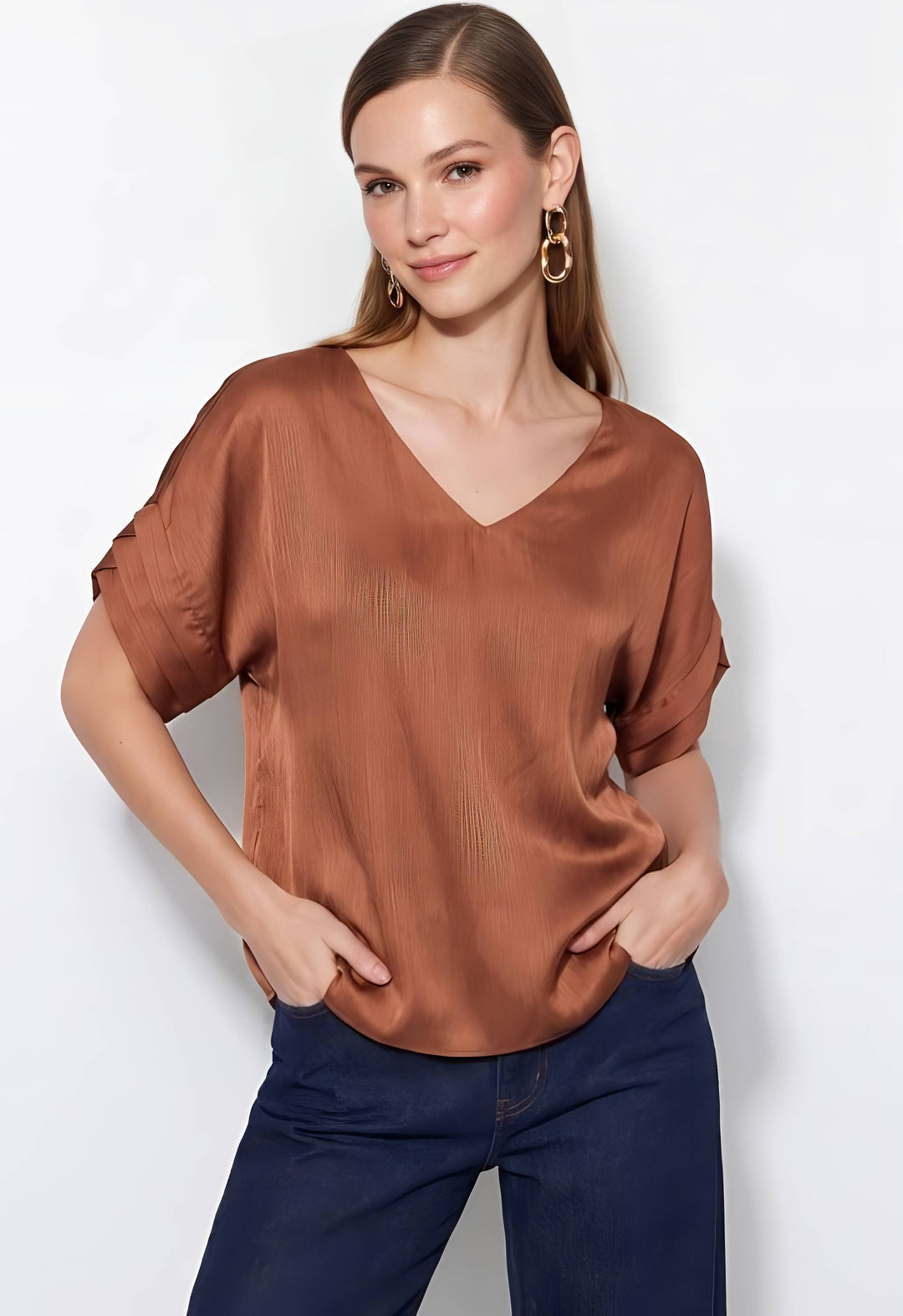 A woman modeling a short-sleeve V-neck satin blouse in a warm rust brown color, paired with dark blue jeans, against a plain white studio background.