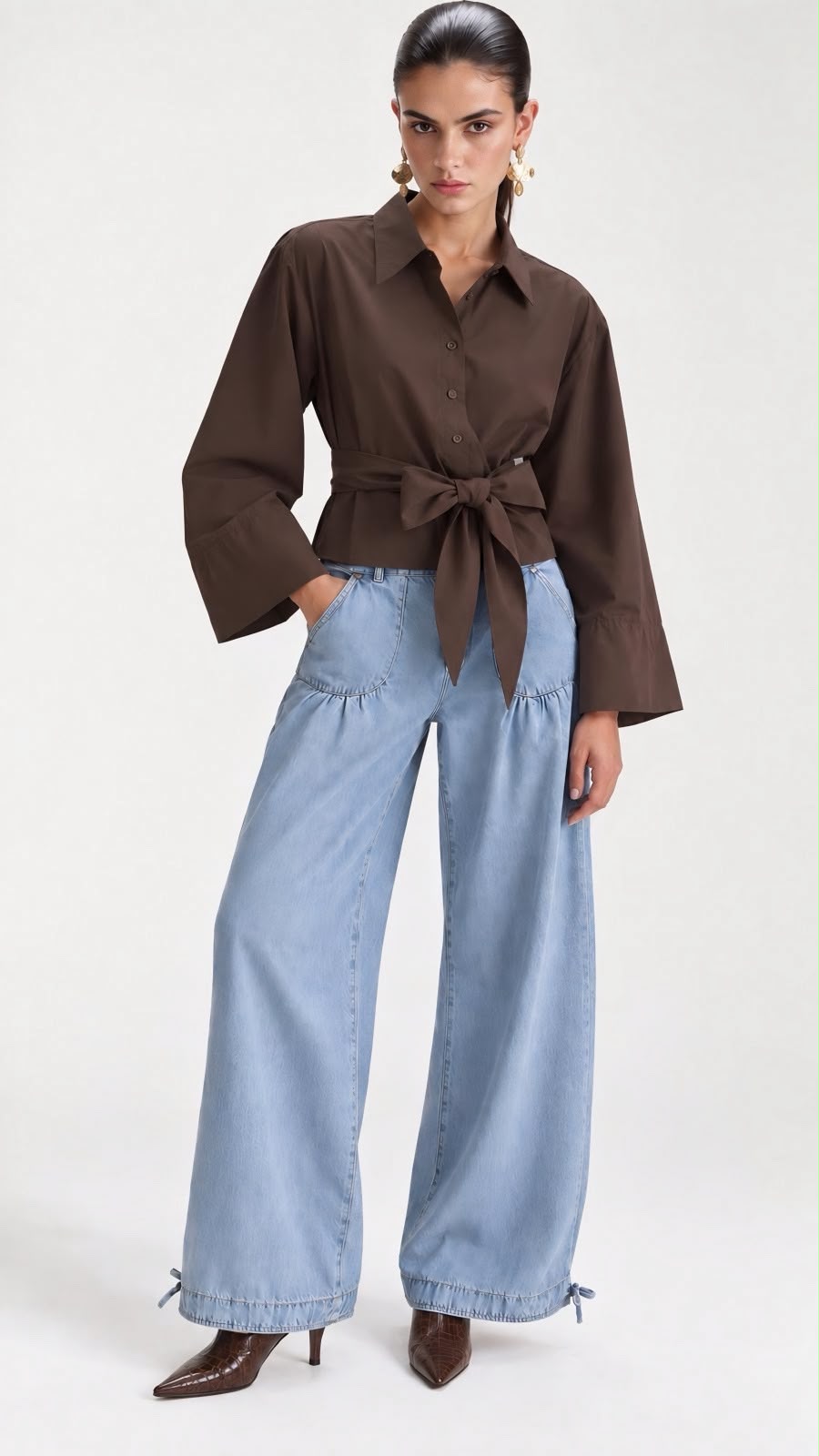 A model wearing a dark brown long-sleeve button-up shirt tied at the waist, paired with wide-leg light blue jeans and brown pointed-toe heels, against a plain light gray studio background.