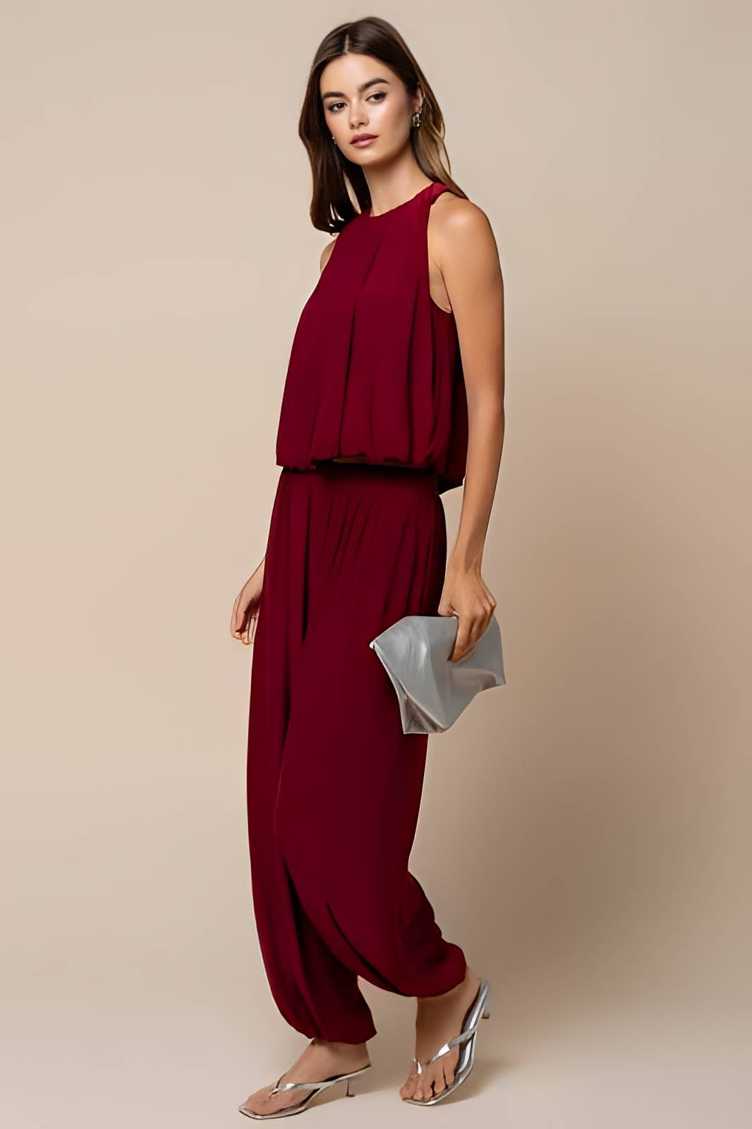 A woman modeling a sleeveless deep burgundy jumpsuit with a draped top and wide-leg pants, holding a metallic silver clutch against a plain beige studio background.