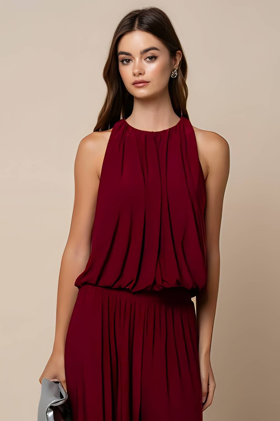A woman modeling a sleeveless, high-neck, pleated jumpsuit in deep burgundy against a solid beige background.