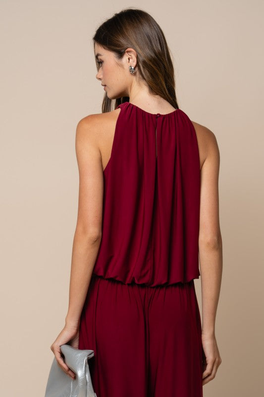 A woman shown from the back wearing a sleeveless deep burgundy jumpsuit with a draped, gathered neckline and matching bottoms, against a plain beige studio background.