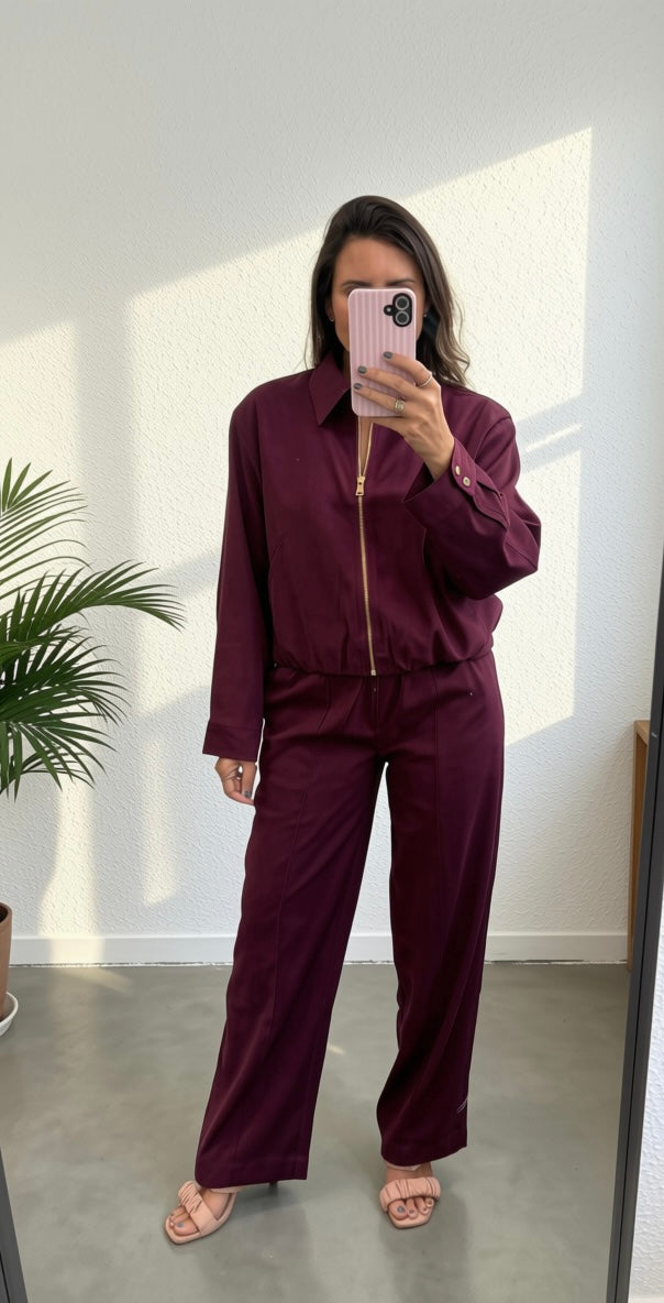 A woman takes a mirror selfie wearing a matching burgundy two-piece tracksuit with a collared long-sleeve top and wide-leg pants, featuring a gold zipper detail.
