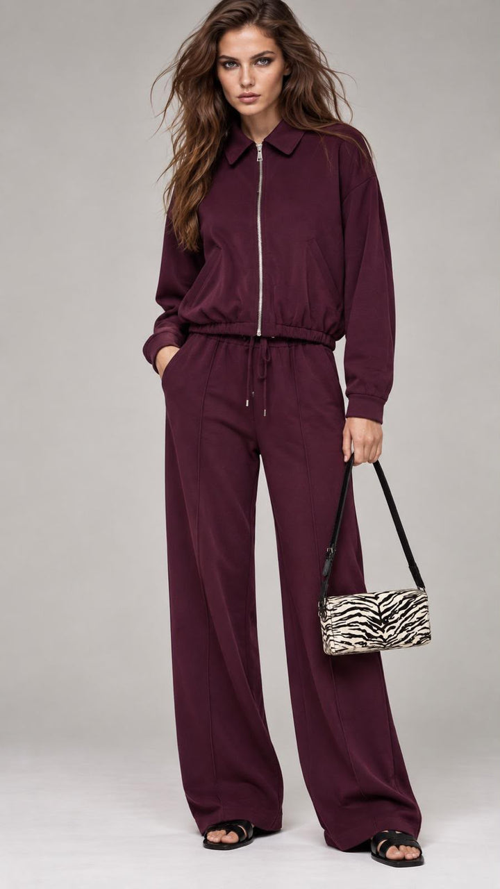 A woman modeling a matching burgundy zip-up jacket and wide-leg pants set, holding a small zebra-print handbag against a light gray studio background.