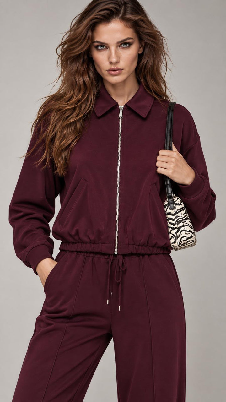 A woman modeling a matching burgundy zip-up jacket and drawstring pants set, holding a black shoulder bag with a zebra print pattern against a neutral gray studio background.