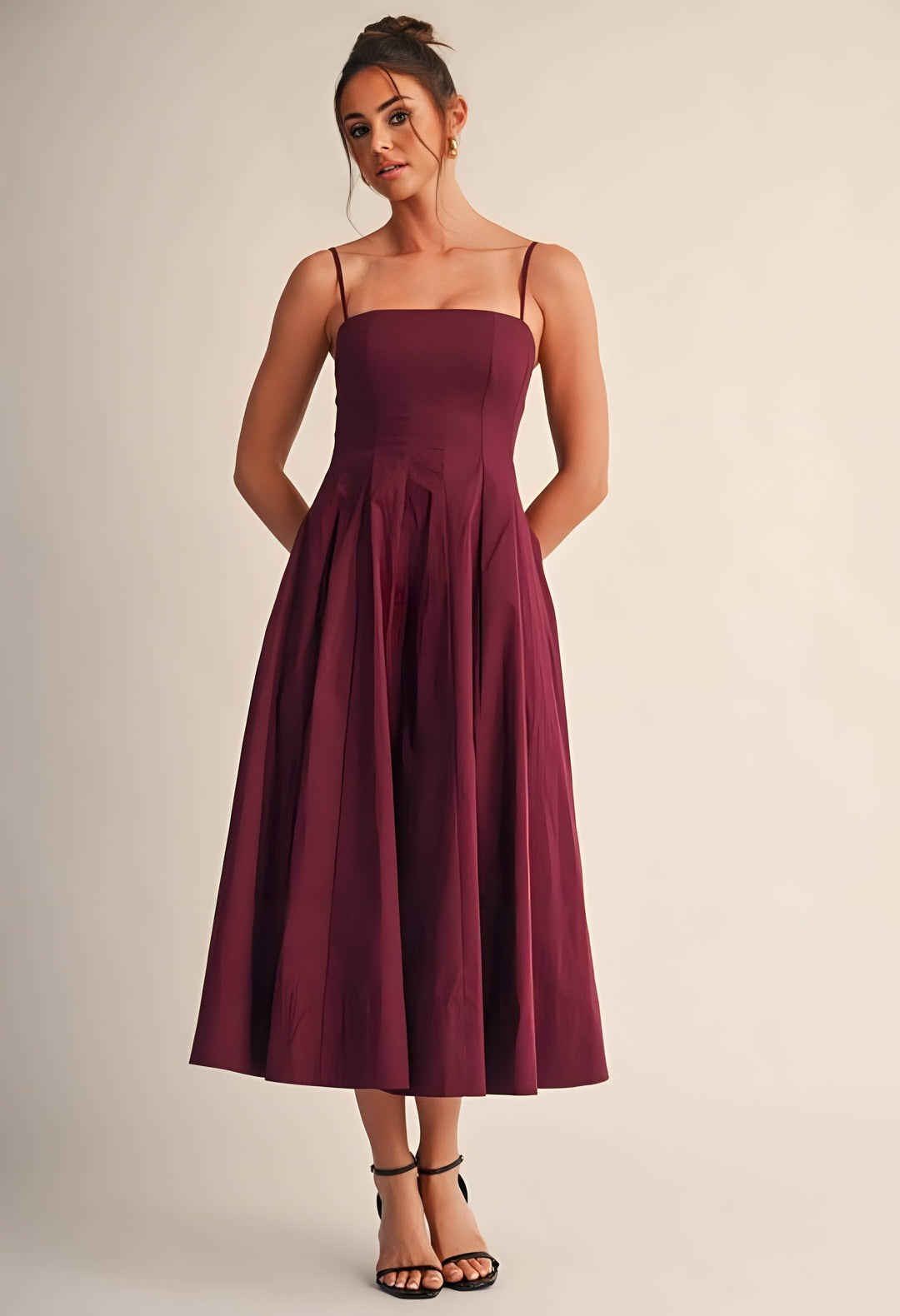 A woman modeling a sleeveless midi dress with thin spaghetti straps, a structured square neckline, a fitted bodice, and a full flared skirt in a deep burgundy color, set against a plain beige studio background.