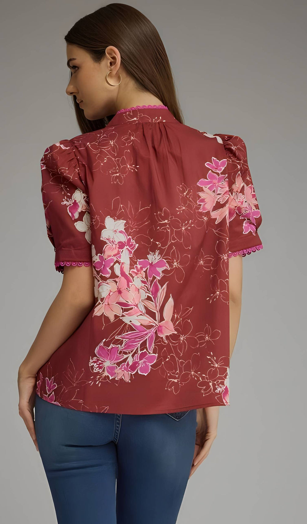 A woman shown from the back wearing a rust-red short-sleeve blouse with a pink and white floral print, paired with blue jeans, against a solid gray studio background.