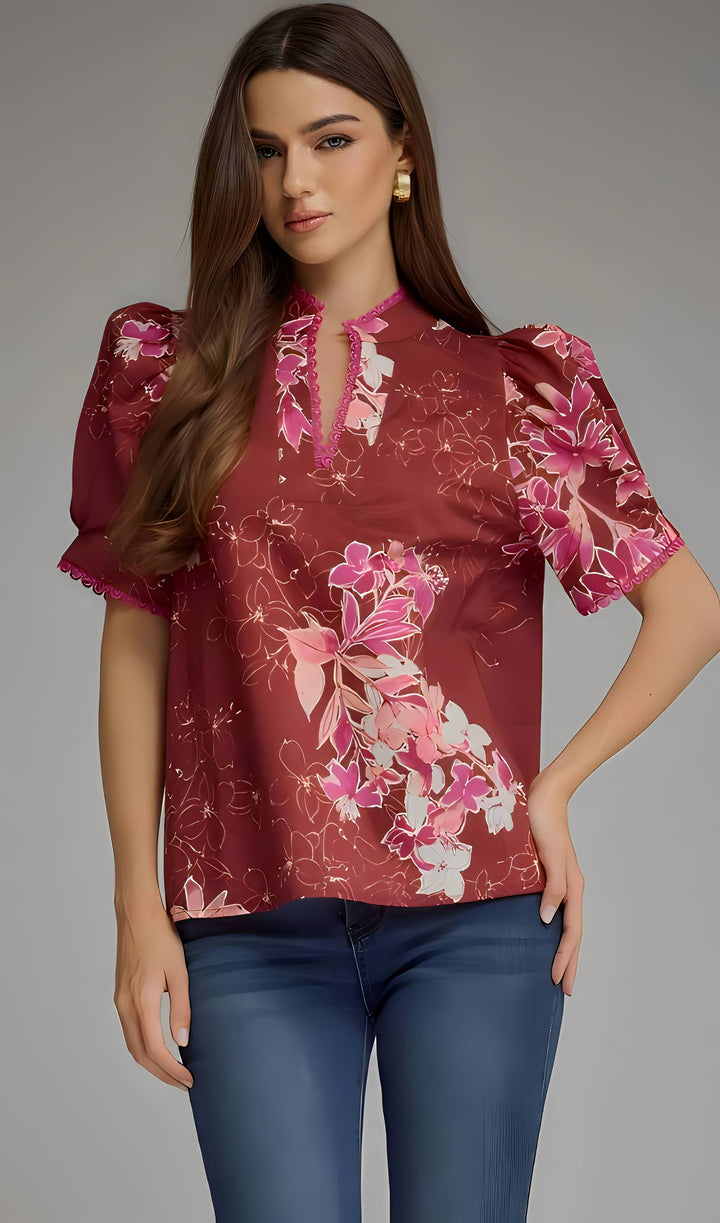 A woman modeling a short-sleeve blouse with puff sleeves, a split neckline, and a pink floral print on a dusty rose background, paired with blue jeans against a gray studio backdrop.