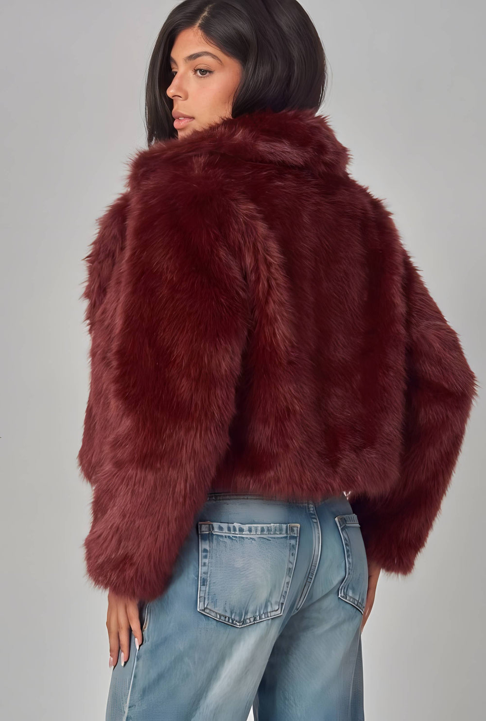 A woman wearing a cropped, plush faux fur jacket in a deep burgundy red, paired with light blue jeans, photographed from the back against a neutral gray studio background.