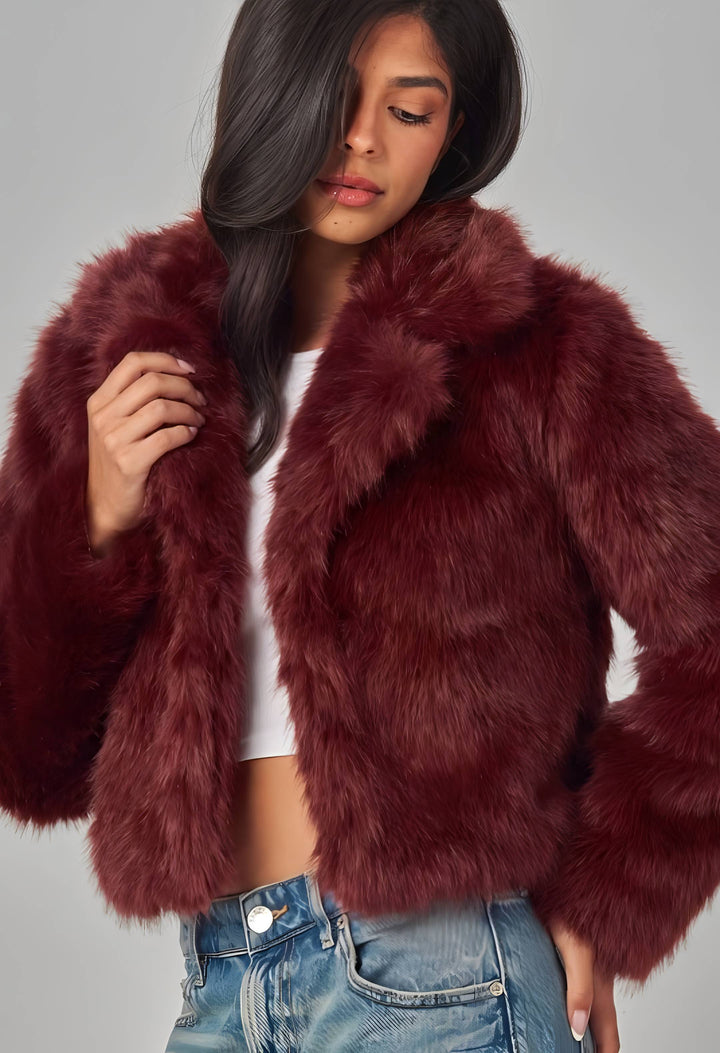 A woman wearing a plush, cropped faux fur jacket in a deep burgundy or maroon shade, styled over a white crop top and blue jeans against a solid gray studio background.